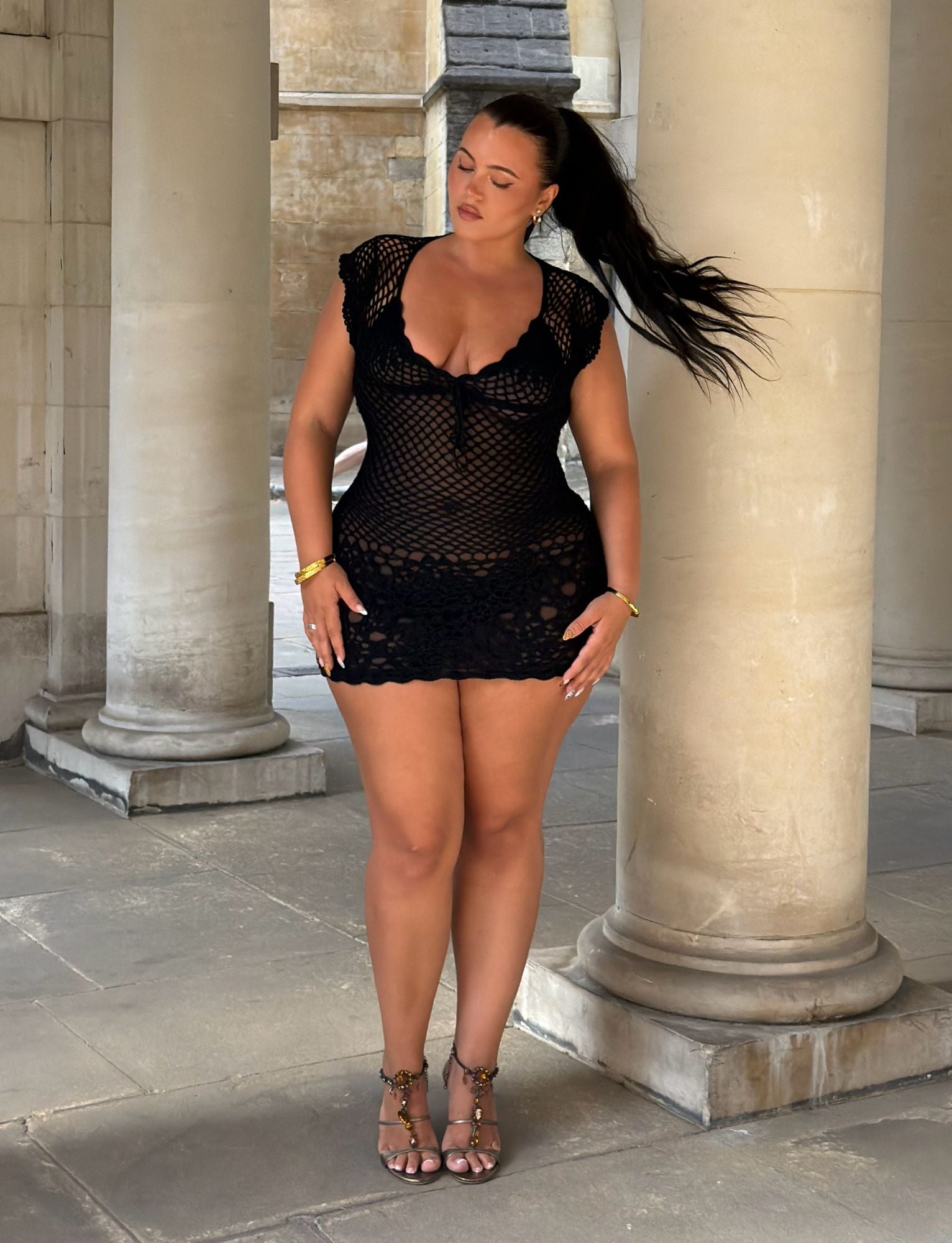 A woman with long dark hair stands between stone columns, wearing the TIGERMIST SASHA CROCHET MINI DRESS in black—a stylish mini dress featuring black crochet fabric and a corset lace-up back—paired with sandals. She looks down, one hand on her hip and the other touching her thigh.