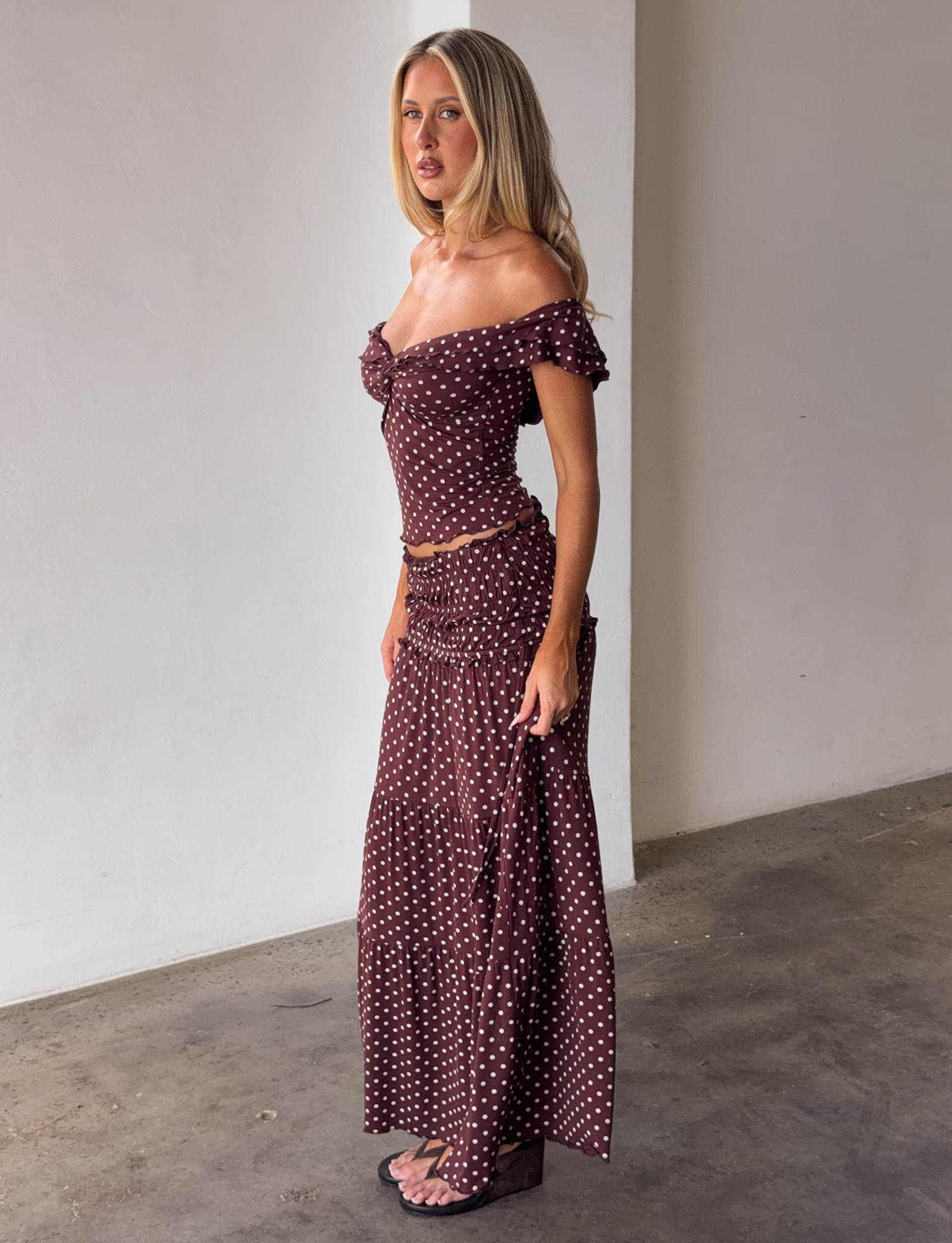 A woman stands indoors against a plain light wall, wearing the TIGERMIST DENALI MAXI SKIRT - BROWN DOT with an off-the-shoulder brown crop top and black wedge sandals.