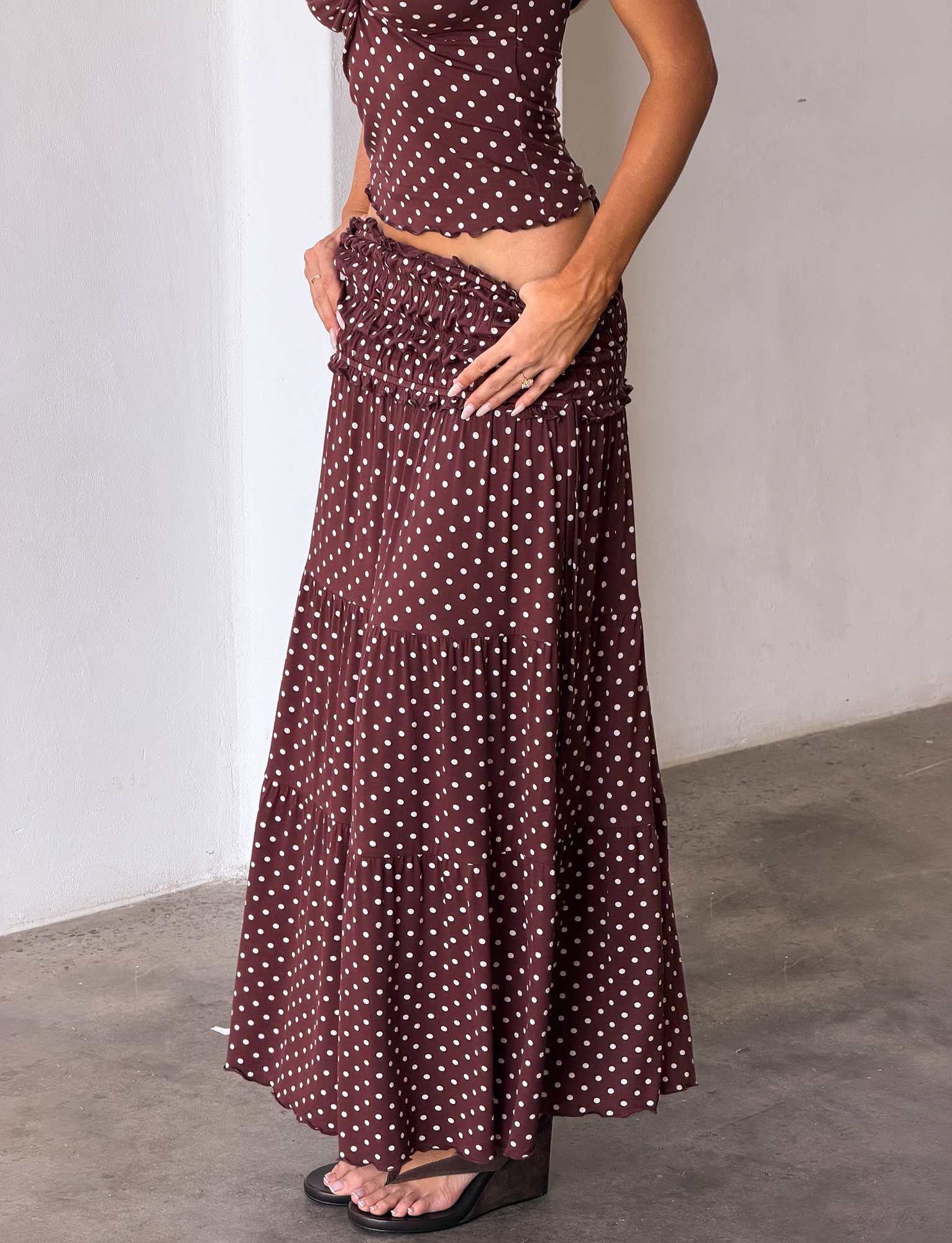 Someone wearing the TIGERMIST DENALI MAXI SKIRT - BROWN DOT with a matching top stands on a concrete floor near a white wall, hands on hips.