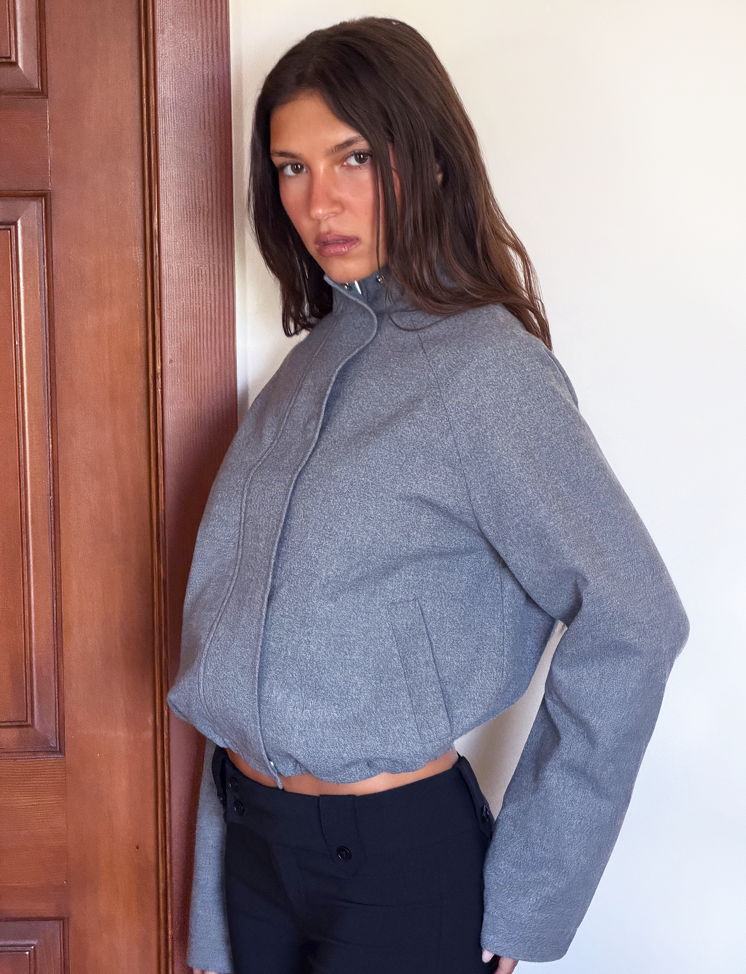 A woman with long brown hair wears the TIGERMIST LENNEX JACKET in heather charcoal, an oversized style with a mock neck collar and zipper deep pockets, paired with black pants. She stands indoors near a wooden door, looking at the camera.