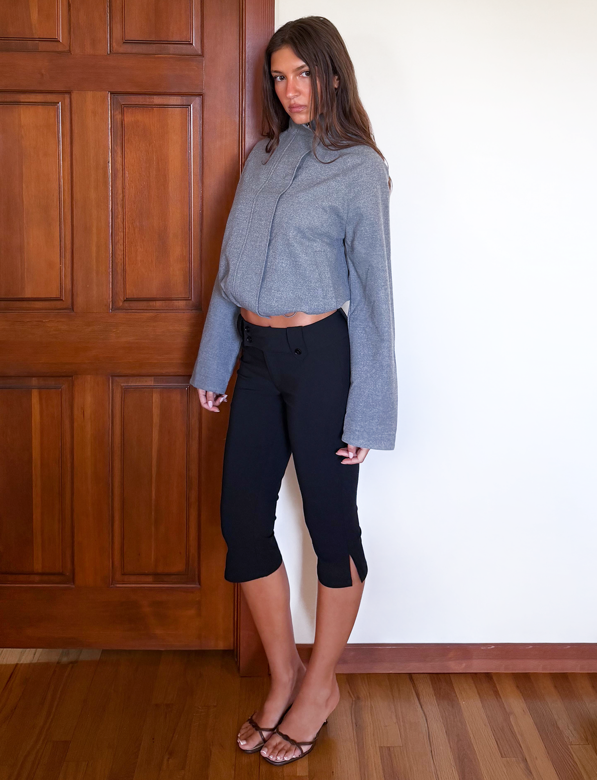 A woman stands indoors by a wooden door, wearing the TIGERMIST LENNEX JACKET in heather charcoal with a mock neck, paired with black capri pants and brown sandals. She has long straight hair and a neutral expression.
