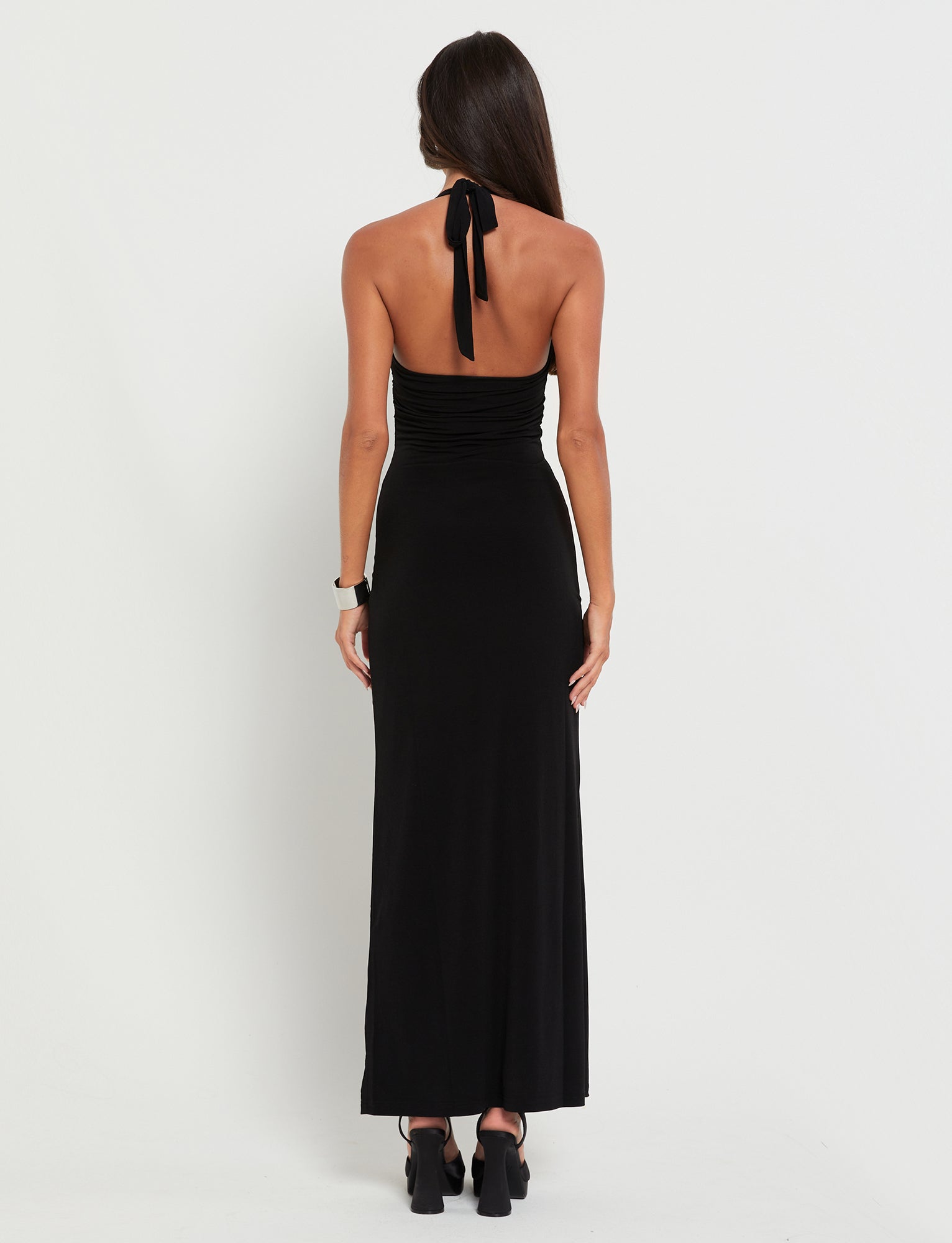 A woman with long brown hair is standing and facing away from the camera, wearing the TIGERMIST CORALINE DRESS - BLACK and black open-toe heels. The halter neck dress ties at the back of her neck and has a fitted silhouette. She has a watch on her right wrist.