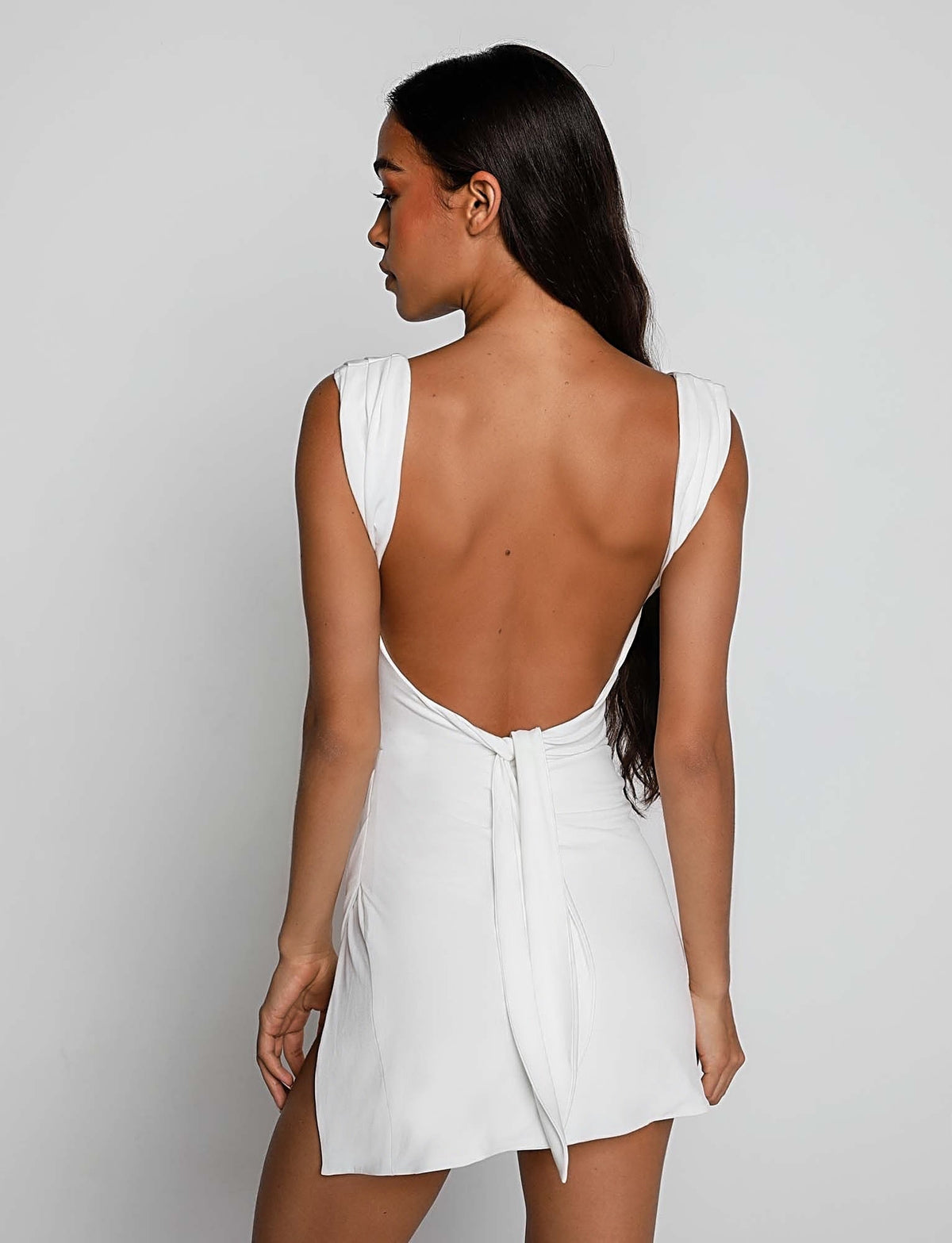 A woman with long, dark hair is shown from the back, wearing the JOSLIN DRESS - WHITE by TIGERMIST. The dress is short, white, and backless with a tie at the lower back. It features wide straps and a relaxed fit. The background is plain and light-colored.
