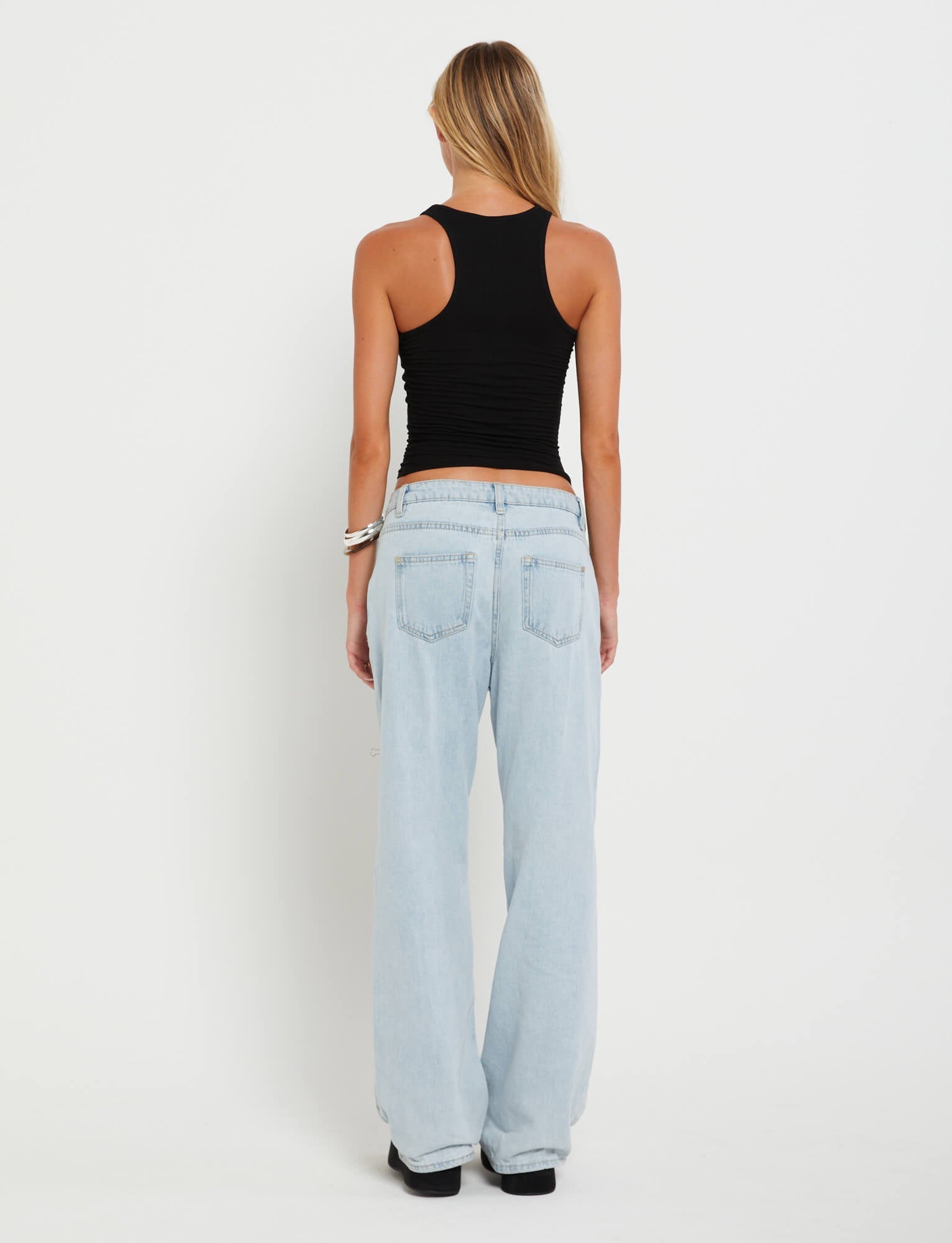 Rear view of a person with long hair wearing a black sleeveless Tiger Mist top and the FELIX JEAN - BLUE : LIGHT DENIM from TIGERMIST. The individual stands against a plain white background, accessorized with a bracelet on their right wrist.