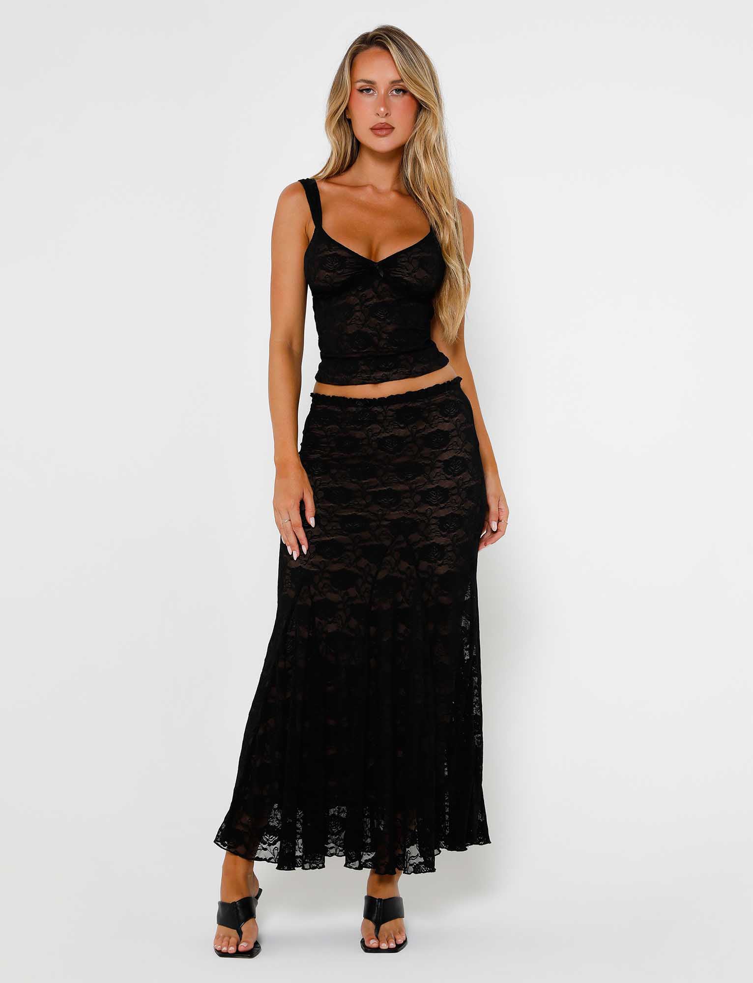 A woman with long blonde hair is wearing the ISADORA SKIRT - BLACK : LACE from TIGERMIST, consisting of a sleeveless top and a long skirt. She is standing against a plain white background, her hands relaxed by her sides, complemented by black heeled sandals.