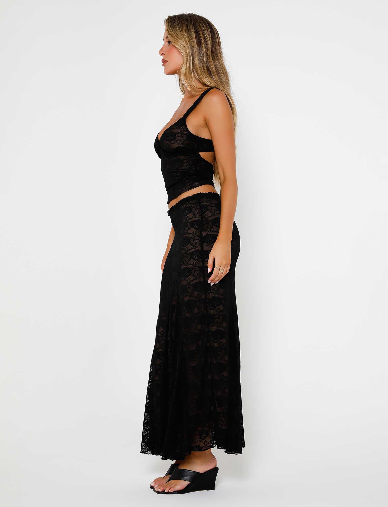 A woman is standing in a side profile against a plain white background, showcasing her chic Tiger Mist ensemble—a black, lace, sleeveless crop top paired with the ISADORA SKIRT - BLACK : LACE from TIGERMIST. Her long blonde hair cascades over her shoulders as she completes the look with stylish black wedge sandals.