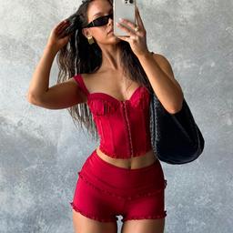 A woman stands against a textured gray background, taking a selfie with her phone. She is wearing a red bustier top and matching shorts, black sunglasses, and a black shoulder bag. One hand is raised to her hair, and she is slightly turned to the side.