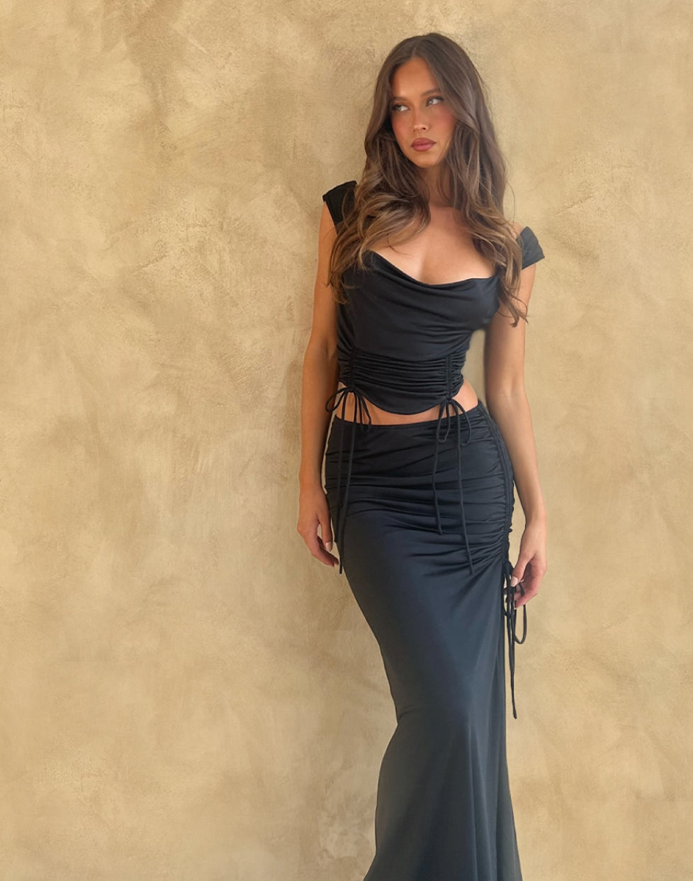 A woman with long wavy hair wears a fitted black dress with a ruched bodice and sides, standing against a beige textured wall and looking to the side.