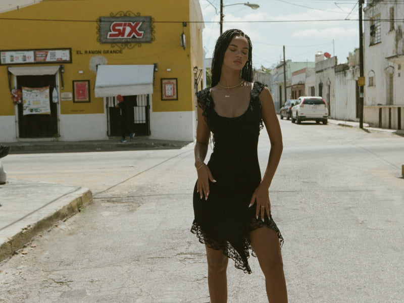 A woman in a black dress stands confidently in the middle of a quiet street with a yellow building and parked cars in the background.