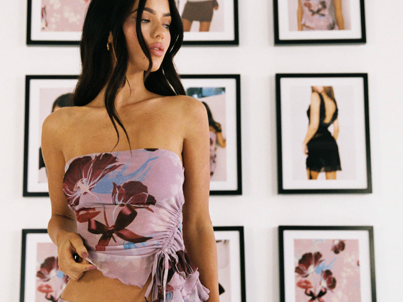A woman wearing a floral strapless top stands in front of a wall decorated with framed fashion photos, some featuring similar floral patterns and dresses.
