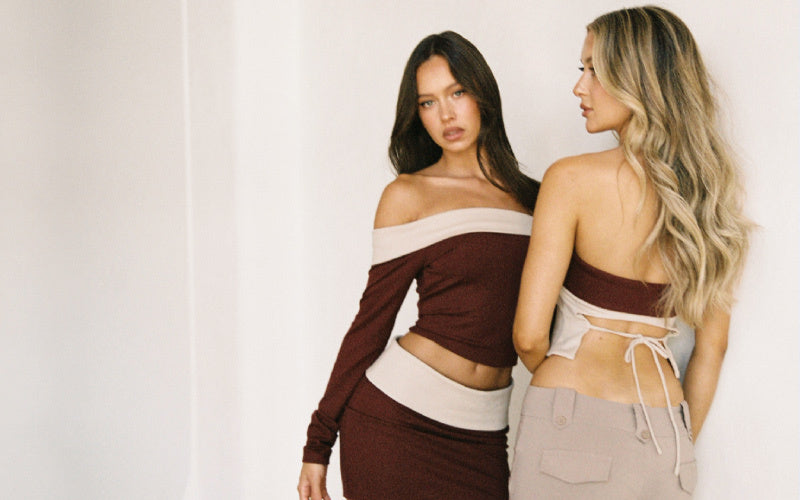 Two women pose together indoors. One faces forward wearing a maroon off-the-shoulder crop top and skirt; the other, with long wavy hair, faces away, wearing a beige backless top and pants.