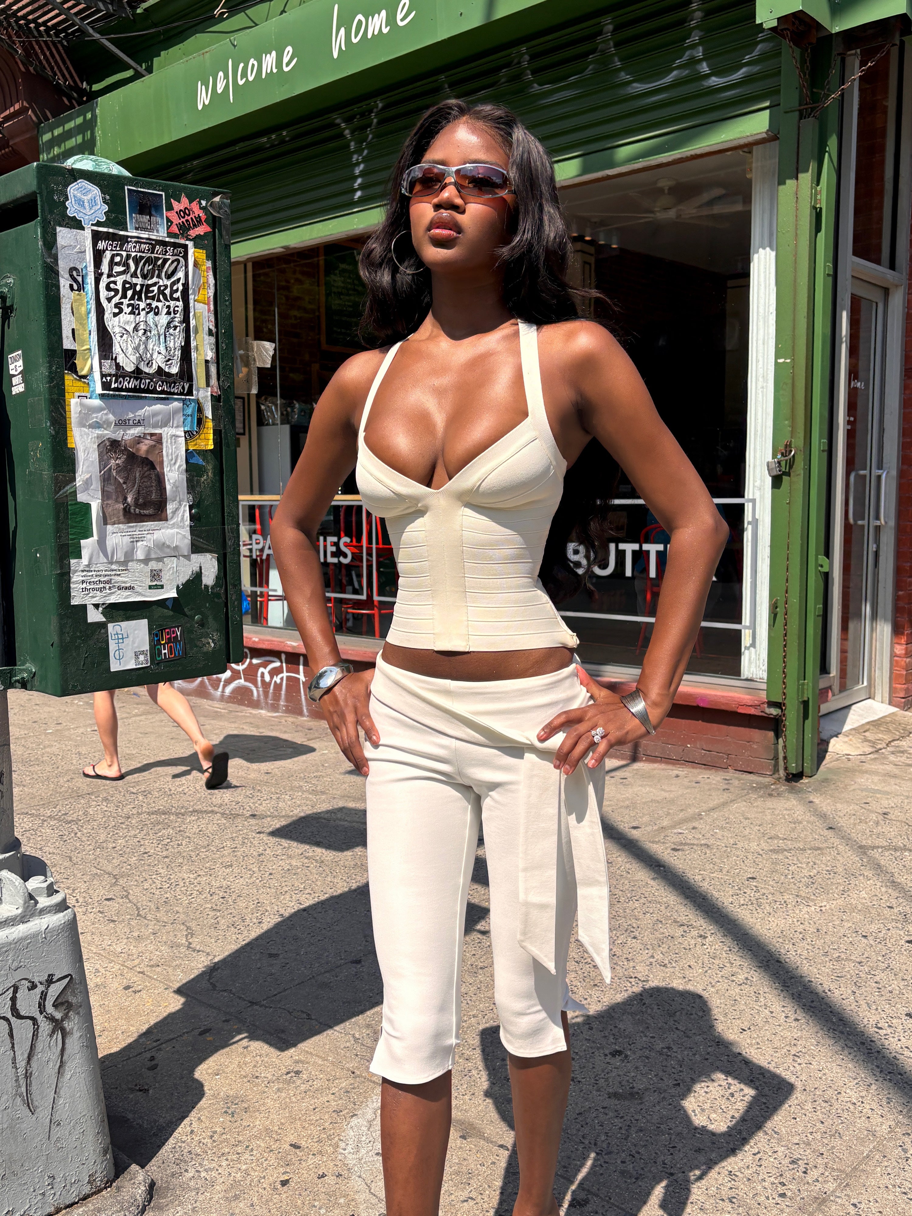 A woman wearing sunglasses, the TIGERMIST SYDEL BANDAGE TOP in cream, and matching cropped pants stands confidently on a sunny city sidewalk in front of a green storefront.