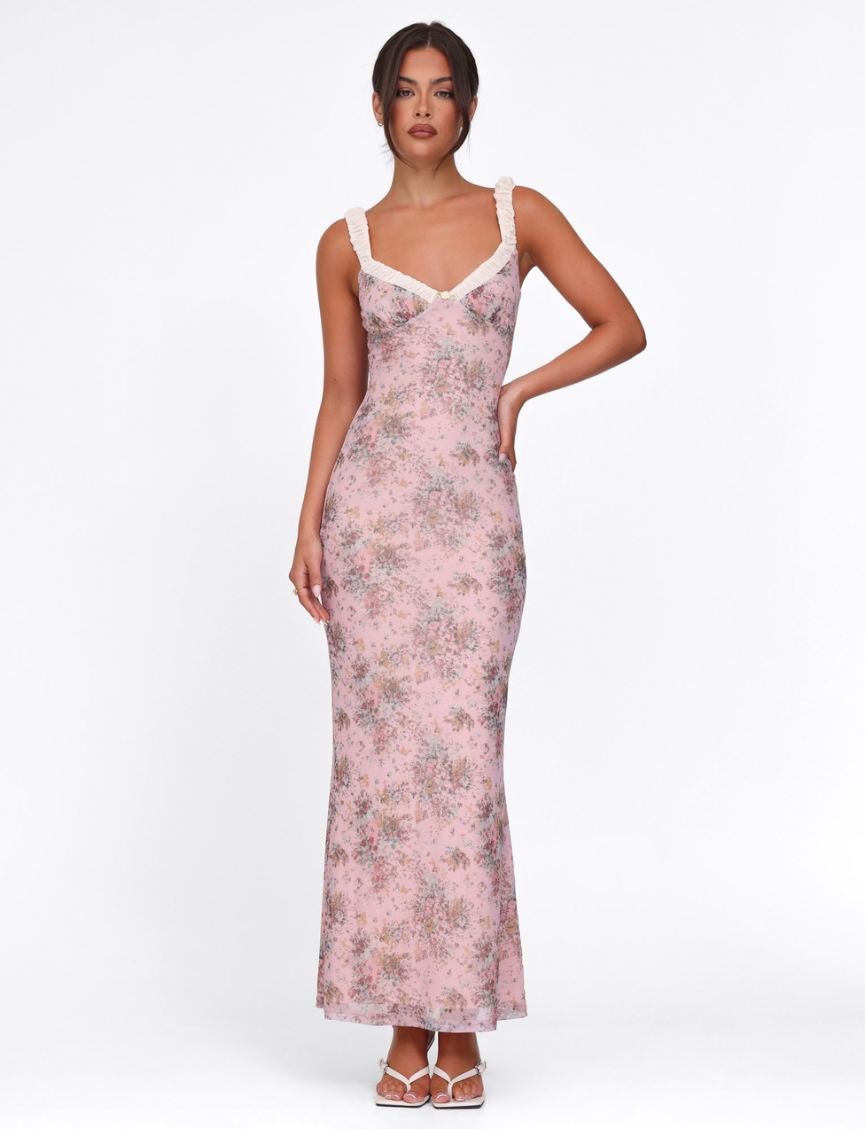 A woman poses against a white background in the TIGERMIST AMARAN DRESS - PINK : CAMO FLORAL, a bodycon maxi with cream lace straps and neckline. She wears her hair up and completes the look with white heeled sandals.