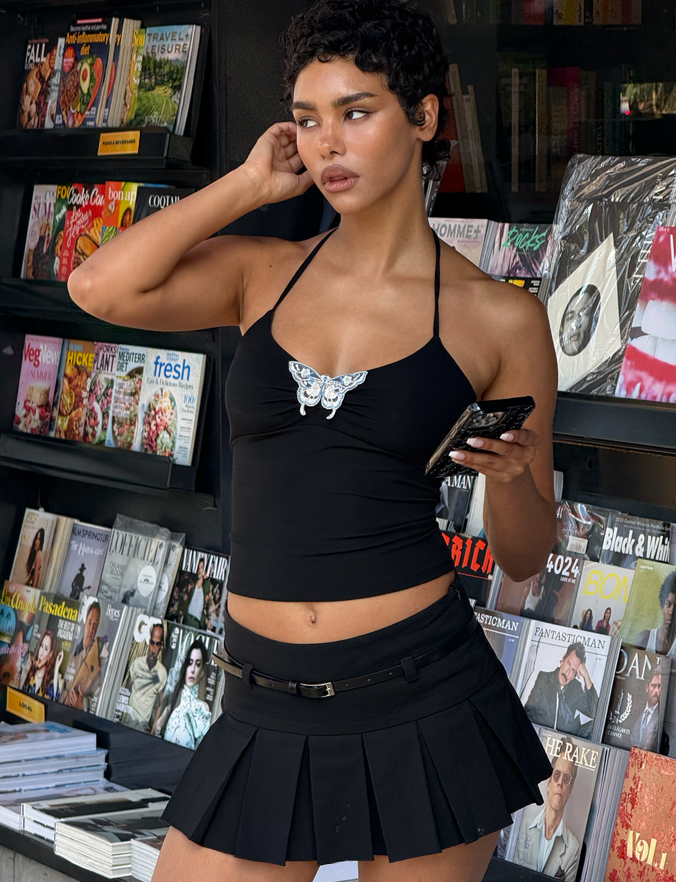 A woman wearing the TIGERMIST ANNELISE SKORT - BLACK, a pleated mini skirt with in-built shorts, and a black halter top with a butterfly design stands by a newsstand, holding her phone as magazines are displayed behind her.