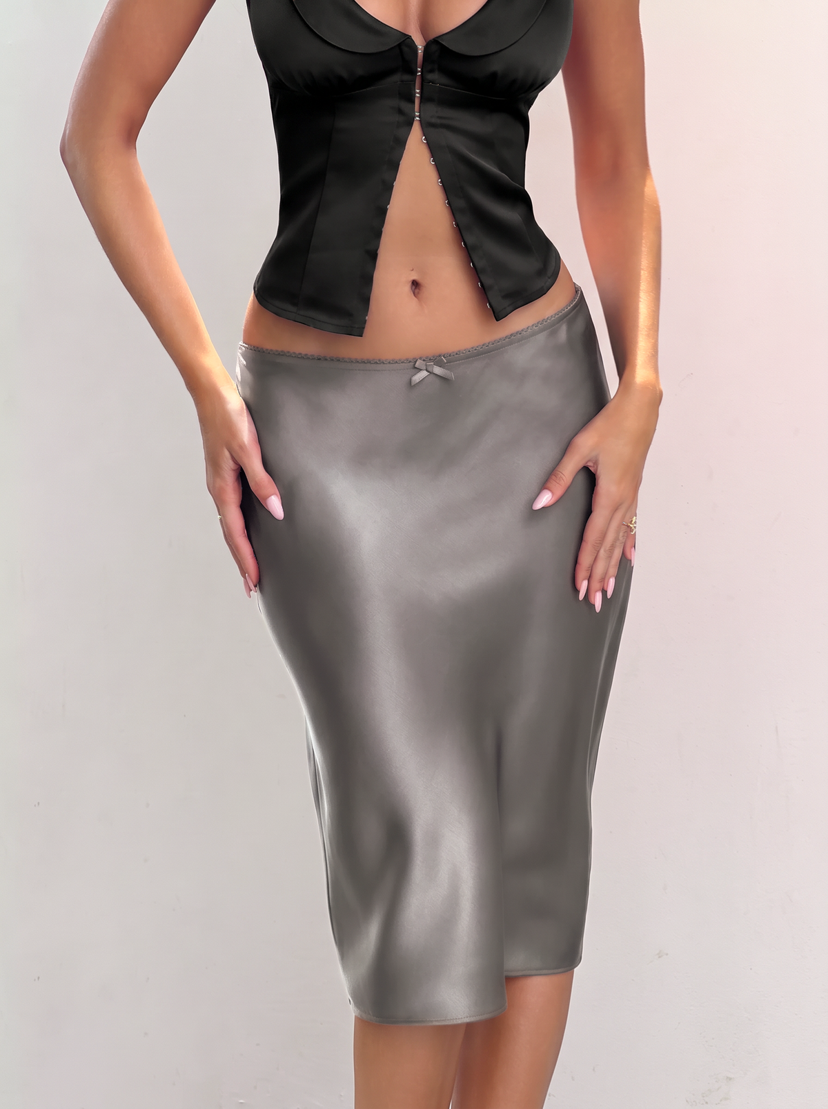 A person wears a charcoal gray ASPEN MIDI SKIRT - CHARCOAL in a 90s column silhouette with a small bow detail at the waistband, paired with a black front-buttoned top.