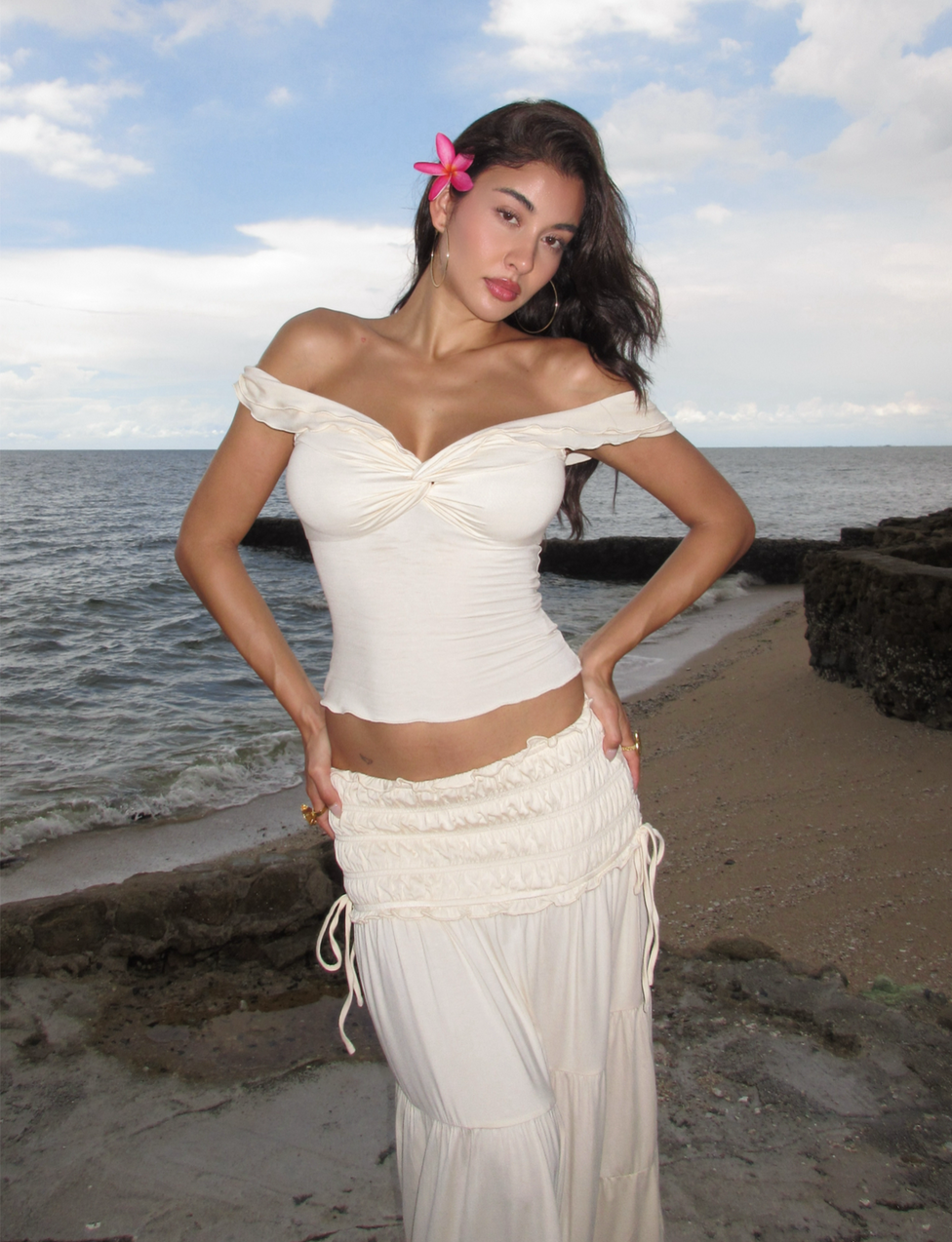 A woman in the TIGERMIST DENALI TOP - NEUTRAL : BONE stands on a sandy beach near the ocean, with a pink flower in her hair and rocks in the background under a partly cloudy sky.