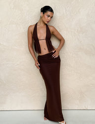 The woman poses elegantly against a textured beige wall, wearing a TIGERMIST ESHA MAXI SKIRT in chocolate brown with an asymmetrical waistband. Her hair is styled neatly, and she pairs the look with white sandals, one hand on her hip and the other gracefully on her leg.