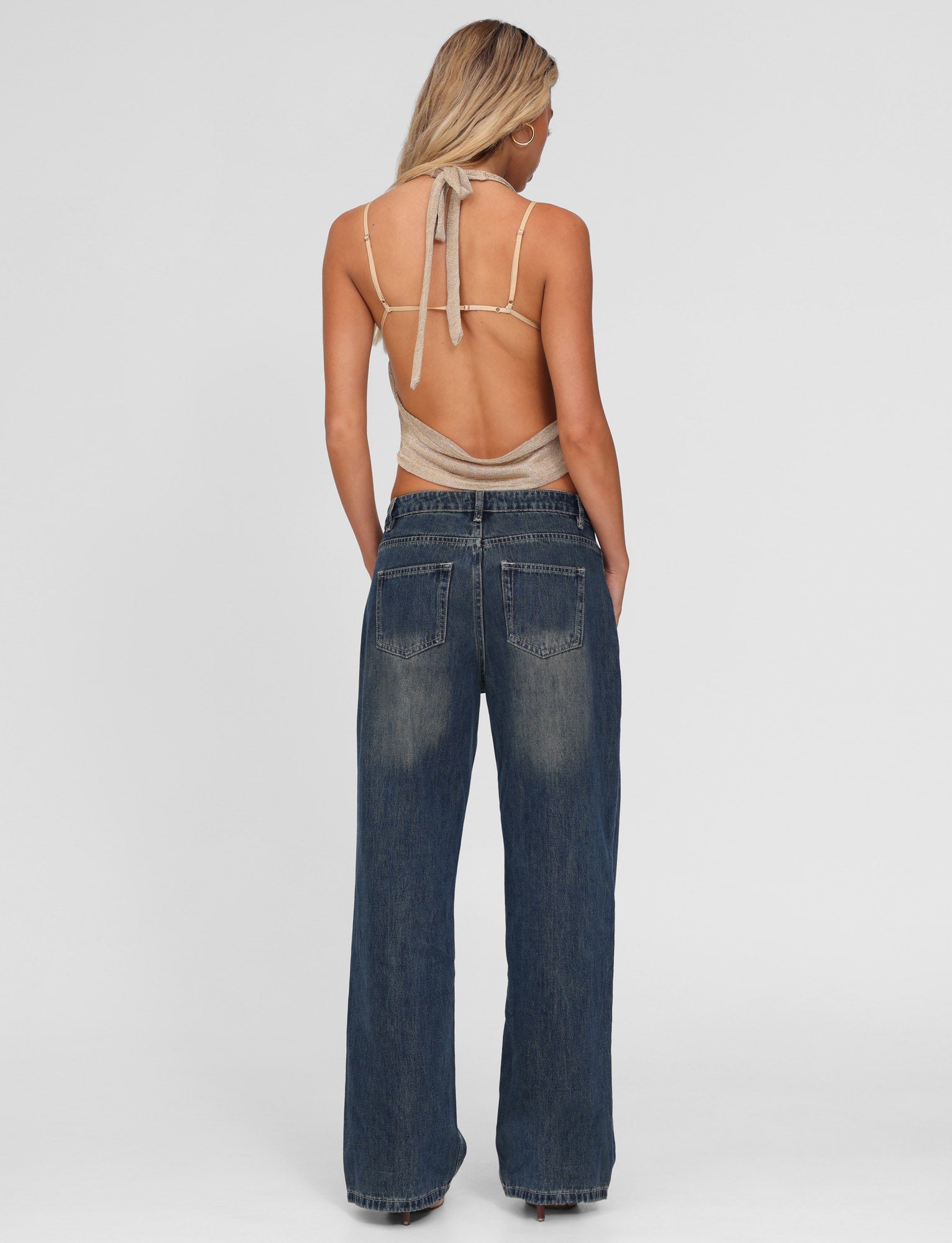 A woman with blonde hair stands facing away, wearing TIGERMIST's FELIX JEAN - BLUE : MID WASH and a beige halter top with an open back. Her hands rest in her pockets as her hair falls over her shoulders.