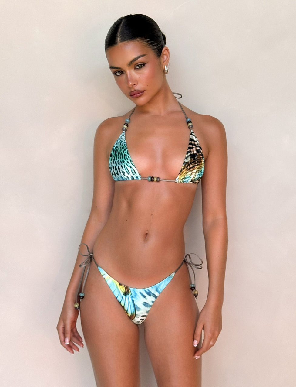A woman poses confidently against a light background, wearing the TIGERMIST FINLEA BIKINI BOTTOM in Butterfly Animal print, showcasing its colorful design and cheeky cut with relaxed arms, slicked-back hair, and neutral makeup.