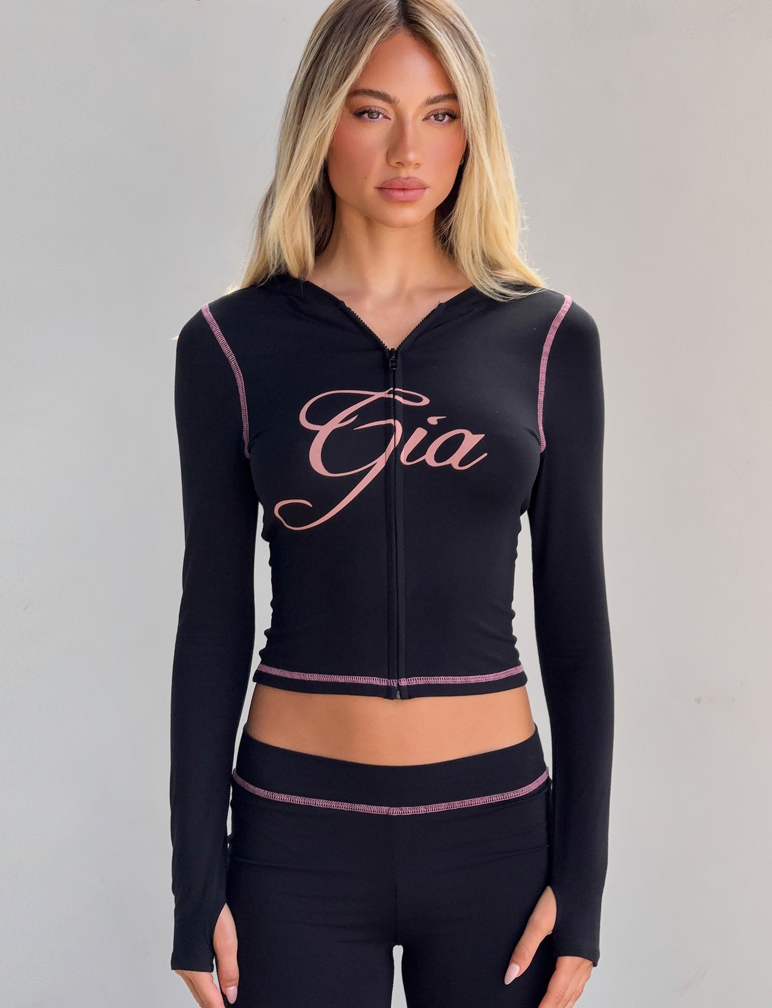 A woman with long blonde hair wears the I.AM.GIA BLARE HOODIE in black, featuring pink edging and "Gia" in bold script across the chest, paired with matching black pants against a plain light background.