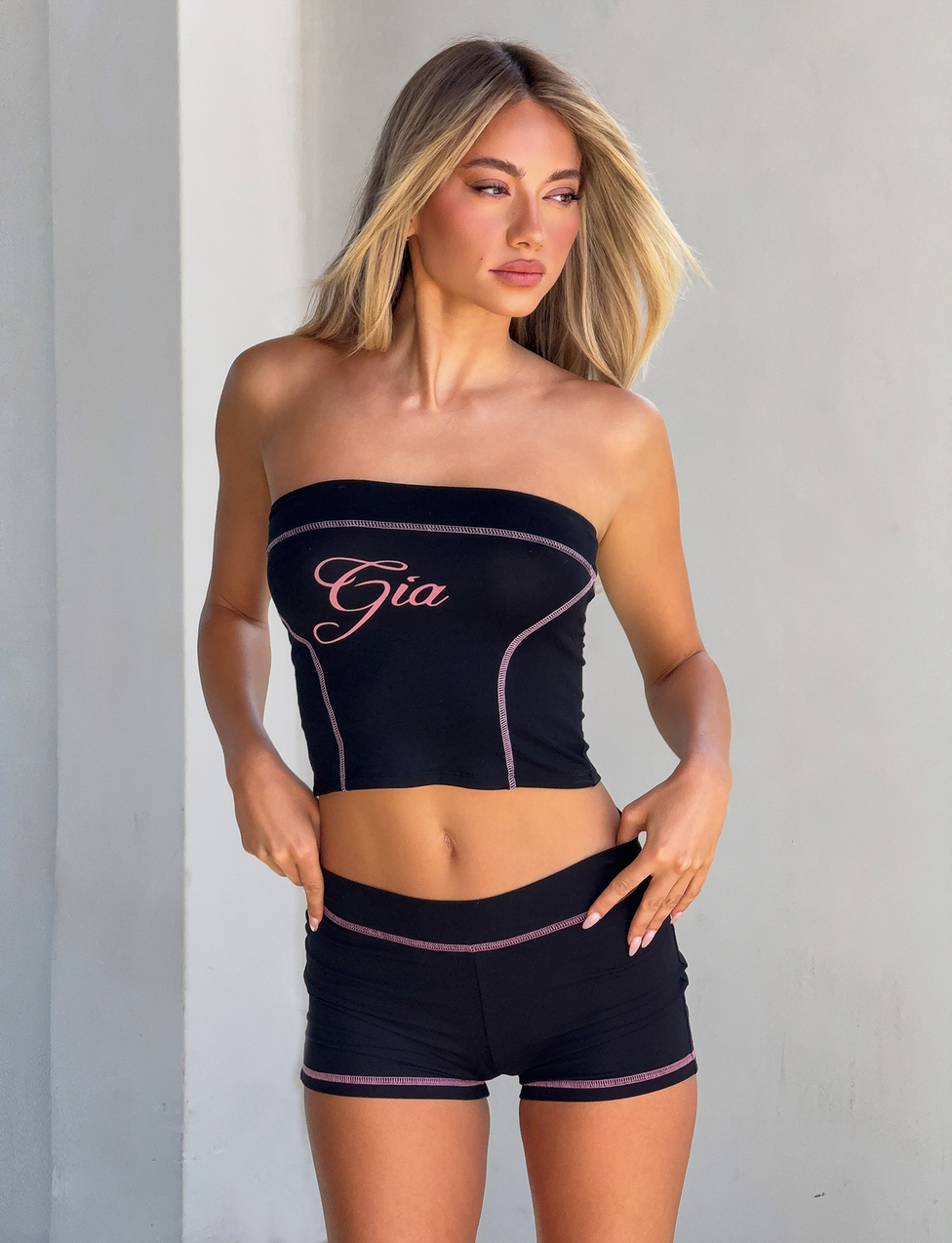 A woman with long blonde hair wears a black "Gia" script tube top and matching I.AM.GIA BLARE SHORT - BLACK, featuring pink stitching and a mid-rise waistband, standing against a light gray wall.