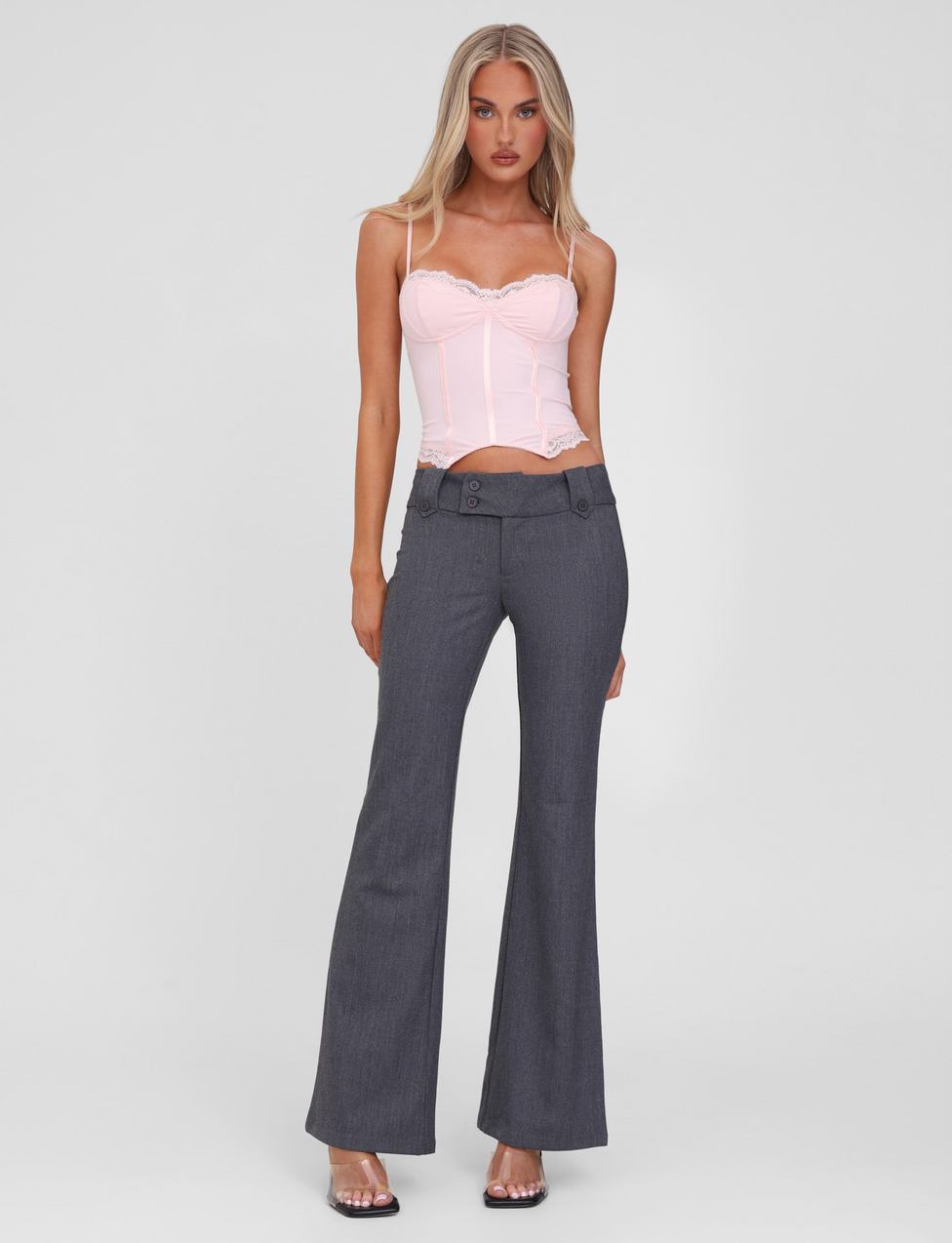 A woman faces forward wearing the TIGERMIST KITTIE PANT - GREY, a high-waisted, flared-leg style with buttons, paired with a light pink corset top and clear-heeled sandals against a plain white background.