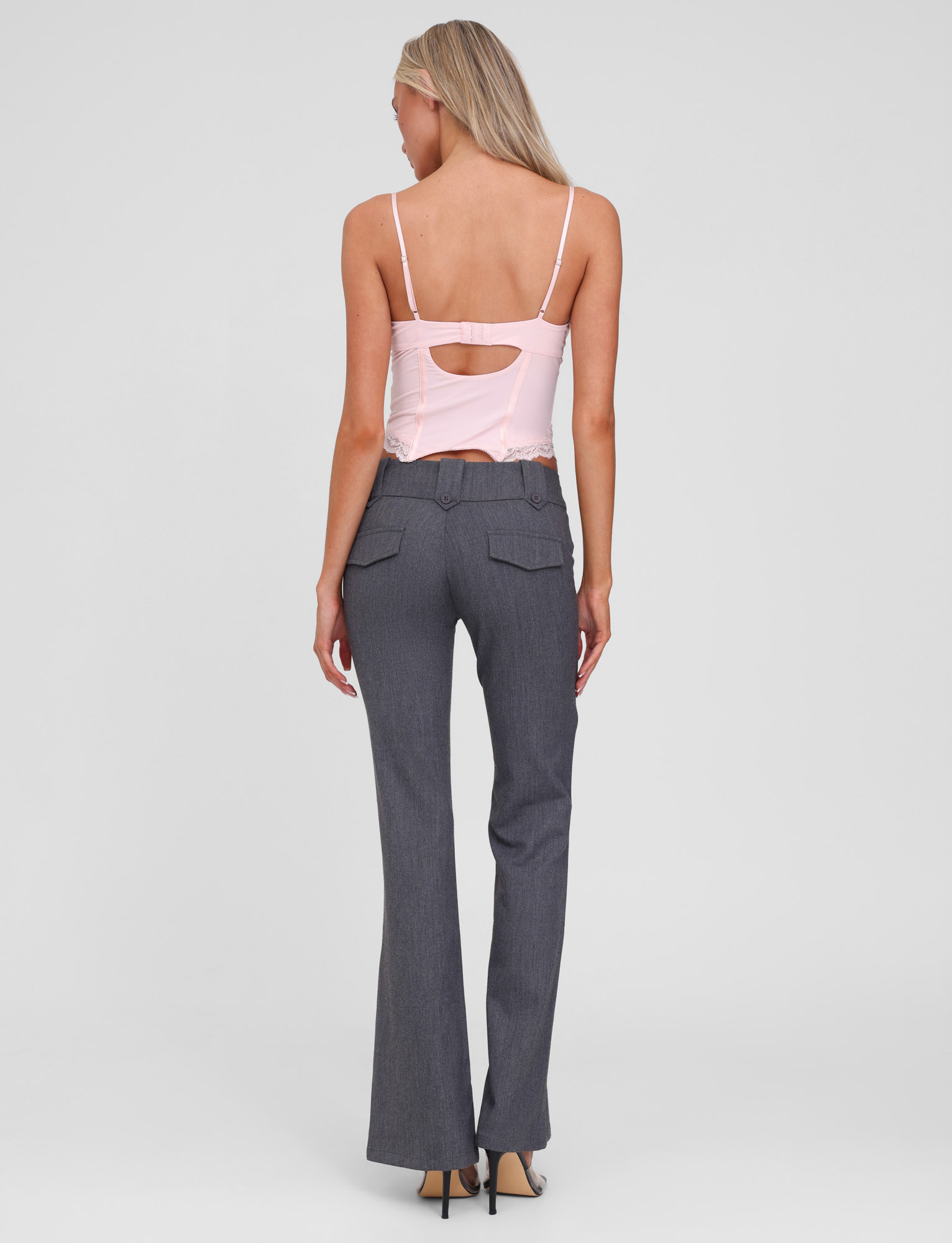 A woman with long blonde hair, wearing a light pink strappy top and TIGERMIST's KITTIE PANT - GREY featuring an asymmetrical waistband, stands facing away against a plain light gray background.