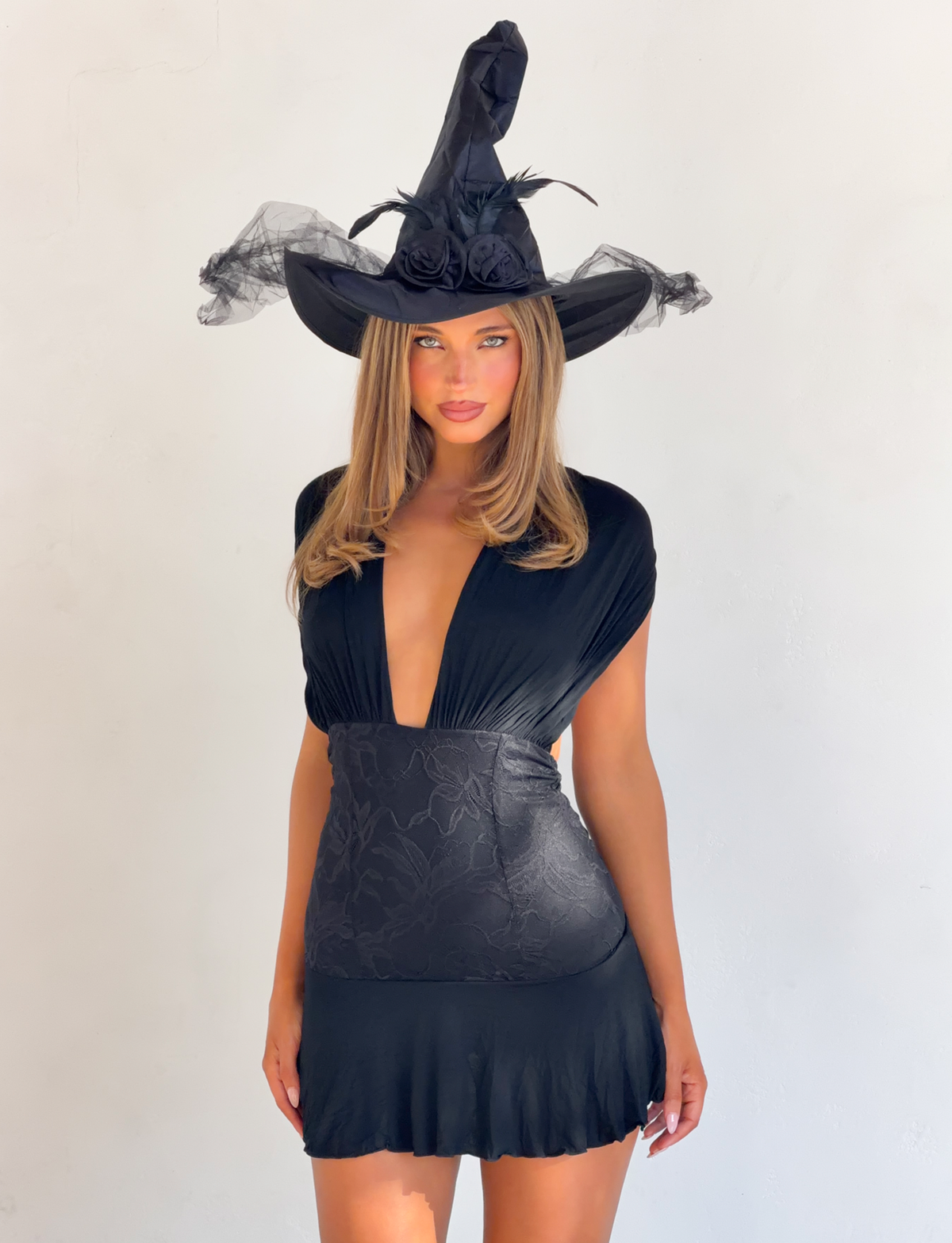 A woman wearing the TIGERMIST CLEO MINI DRESS - BLACK with a deep V-neck, lace waist, and a tall pointed hat with black flowers stands against a plain white background.