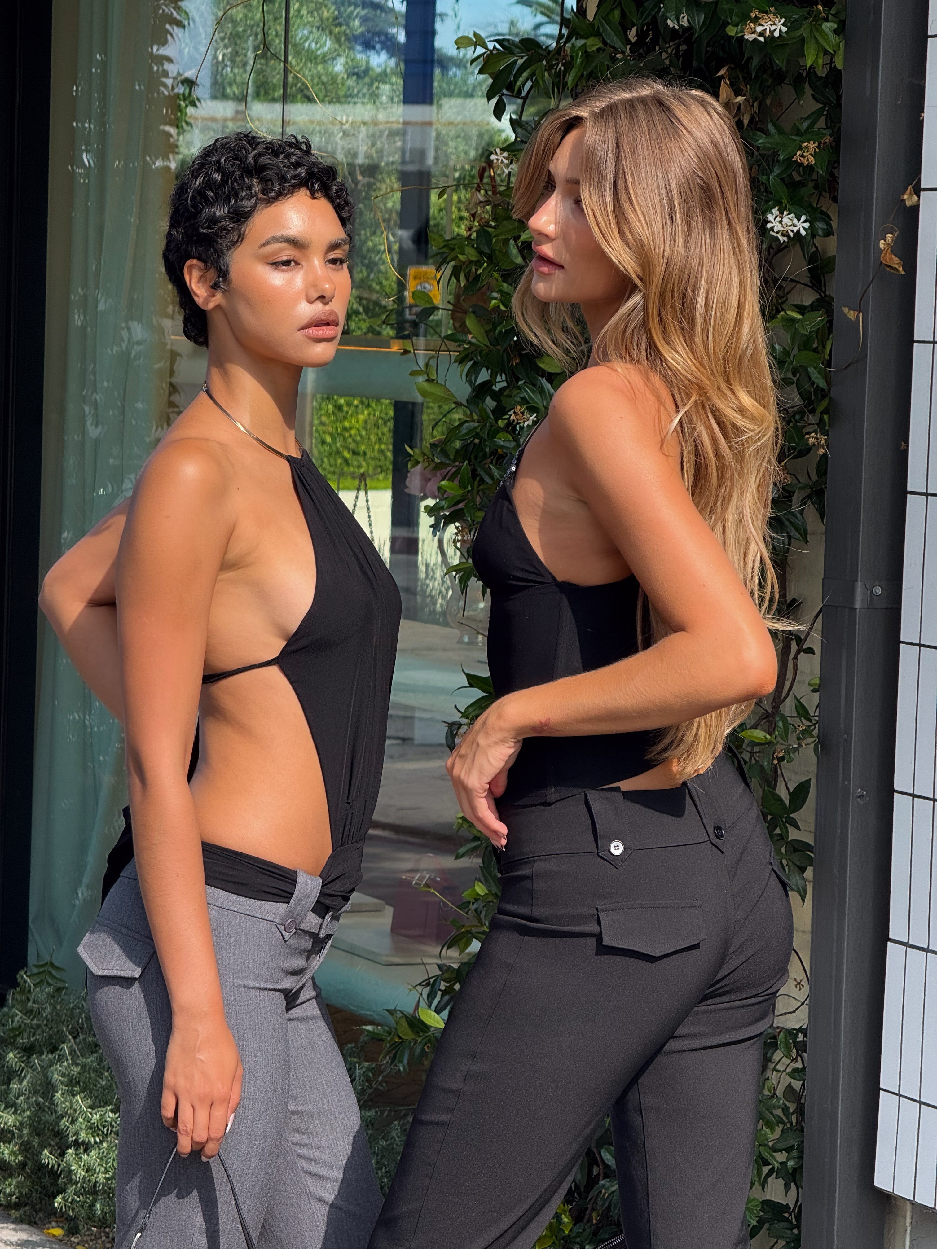 Two women stand outdoors in front of greenery. One wears the TIGERMIST FENELLA TOP - BLACK with high-waisted pants, while the other pairs a sleeveless black sequin trim tank with gray pants.