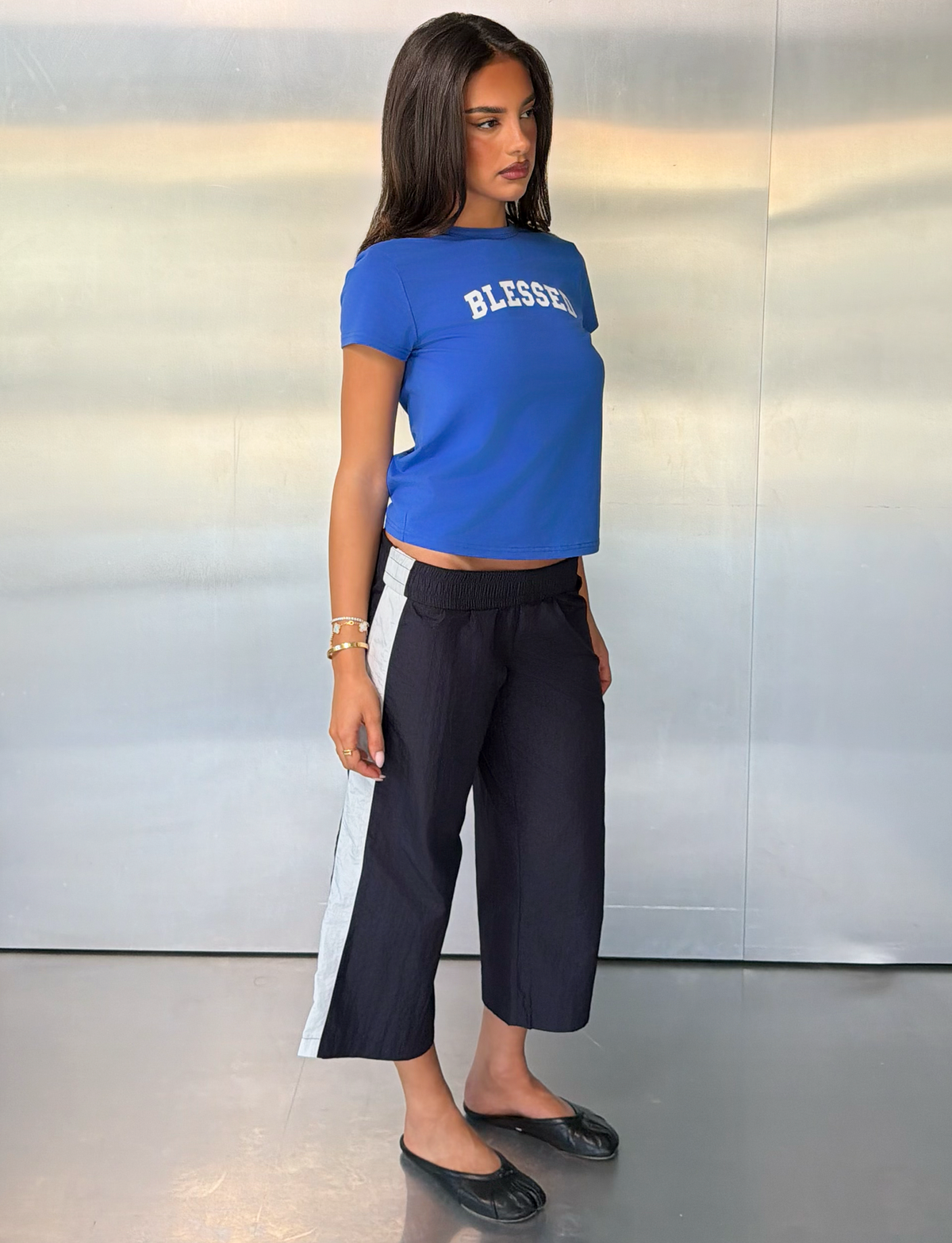 A woman stands sideways against a metallic wall, wearing the TIGERMIST BLESSED BABY TEE in blue—a vintage relaxed fit graphic top—paired with black wide-leg pants featuring white side stripes, black flats, and bracelets. She looks slightly over her shoulder.