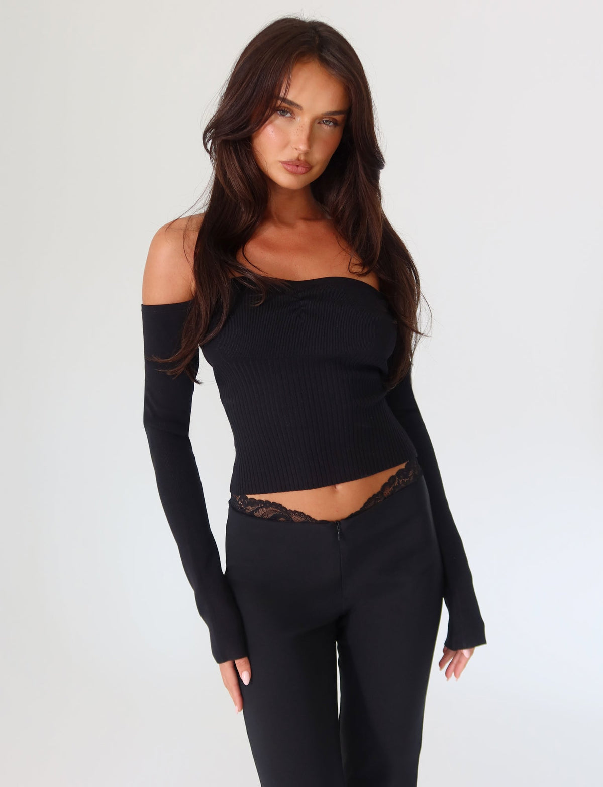 A woman with long hair is elegantly posed against a plain white background, showcasing the TIGERMIST LILYANNA LONG SLEEVE TOP in black. She wears coordinating black pants accentuated by lace detail at the waist, exuding sophistication with her arms gracefully resting at her sides in this fitted, off-the-shoulder ensemble.