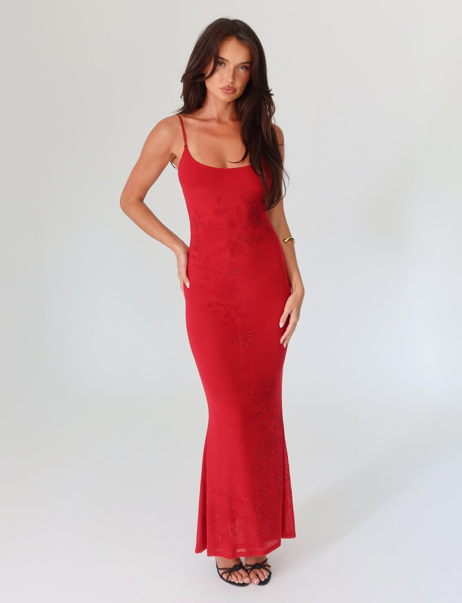 A woman elegantly posed in the Cherry Red Octavia Maxi Dress by Tigermist stands against a plain white background. Her long hair and black strappy heels complement the floral-embellished, sleeveless design, as she confidently rests her left hand on her hip.