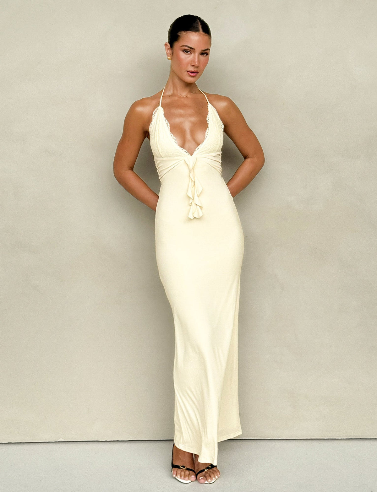 A person stands against a neutral background, wearing the DEENA MAXI DRESS - YELLOW : LEMON from TIGERMIST—a long, form-fitting yellow dress with delicate lace bust cups, a deep V-neck, and halter straps. Their hair is styled in a sleek updo, and they wear open-toed heels. Their arms are relaxed at their sides.