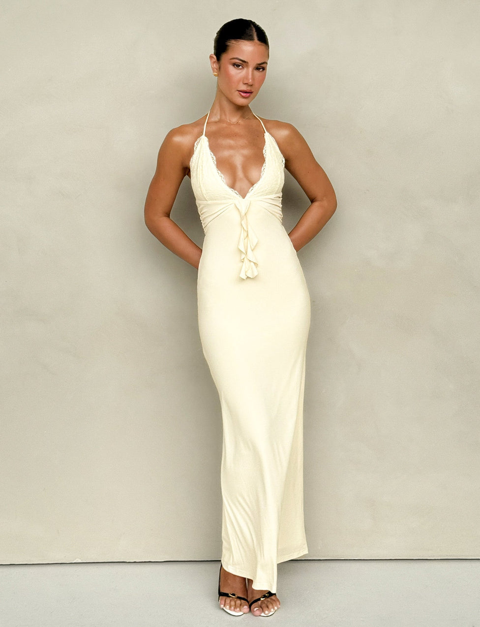 A person stands against a neutral background, wearing the DEENA MAXI DRESS - YELLOW : LEMON from TIGERMIST—a long, form-fitting yellow dress with delicate lace bust cups, a deep V-neck, and halter straps. Their hair is styled in a sleek updo, and they wear open-toed heels. Their arms are relaxed at their sides.