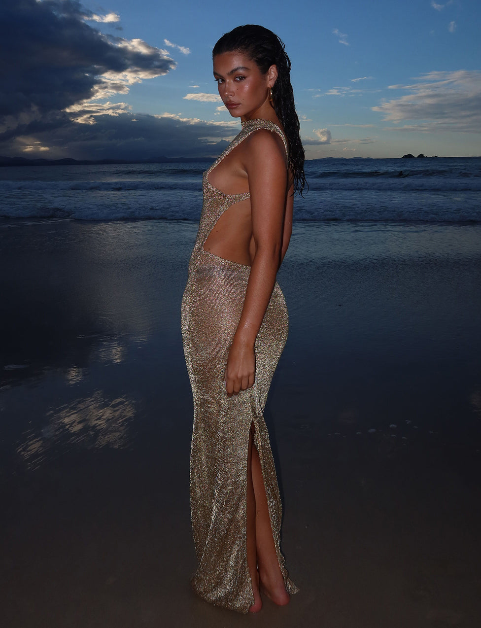 A woman stands on a beach at sunset, wearing the dazzling ZAELA DRESS - GOLD from TIGERMIST. The sky behind her is filled with dark clouds, and the ocean is calm with gentle waves lapping at the shore. The golden dress reflects the fading light, highlighting her silhouette.