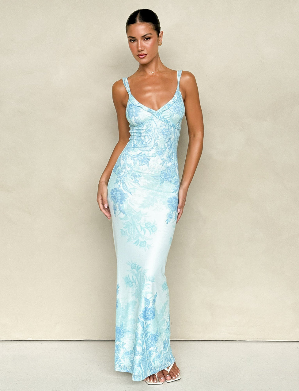 A person wearing the GUILIANNA FRILL MAXI DRESS - BLUE : FLORAL : TAPESTRY from TIGERMIST poses against a plain beige background. They have long hair tied back and are standing with one hand on their hip, highlighting the frill shoulder strap, while wearing white sandals.