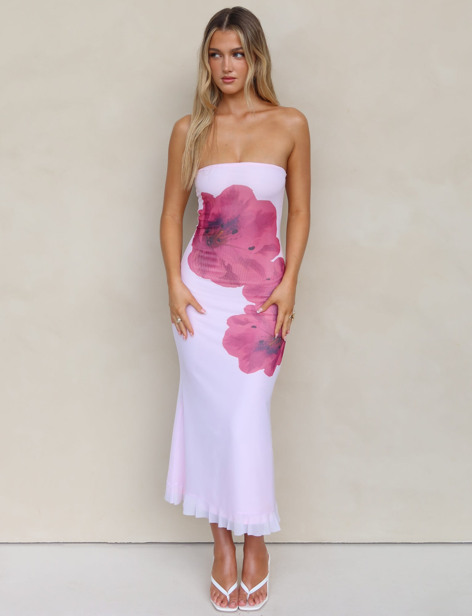 A woman with long blonde hair is wearing the LILLIAN DRESS - PINK by TIGERMIST, a strapless, form-fitting white midi dress featuring large pink floral designs. She poses against a plain beige background and completes the look with white open-toe heels.