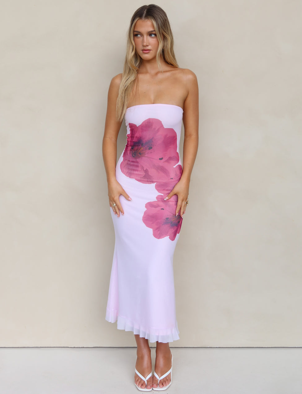 A woman with long blonde hair is wearing the LILLIAN DRESS - PINK by TIGERMIST, a strapless, form-fitting white midi dress featuring large pink floral designs. She poses against a plain beige background and completes the look with white open-toe heels.