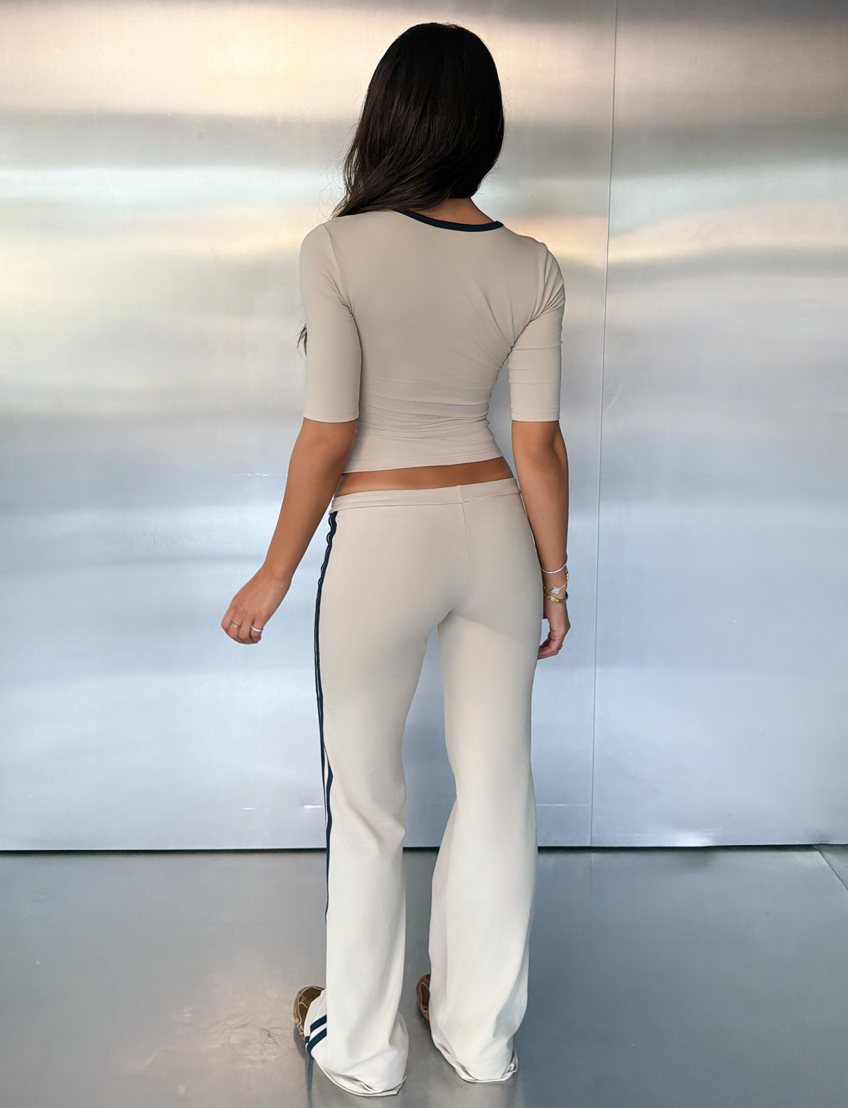A woman with long hair stands facing away, wearing the TIGERMIST SHANICE TOP in Sand—a fitted, light gray three-quarter sleeve top—paired with matching flared pants featuring navy side stripes, in a minimalist room with metallic walls and floor.