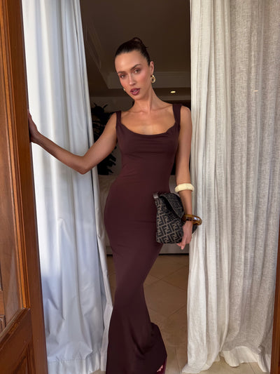 A woman stands in a doorway, wearing the TIGERMIST MORANA DRESS - BROWN : CHOCOLATE. She holds a patterned handbag, wears large bangles on one arm, her hair pulled back, and is framed by white curtains.