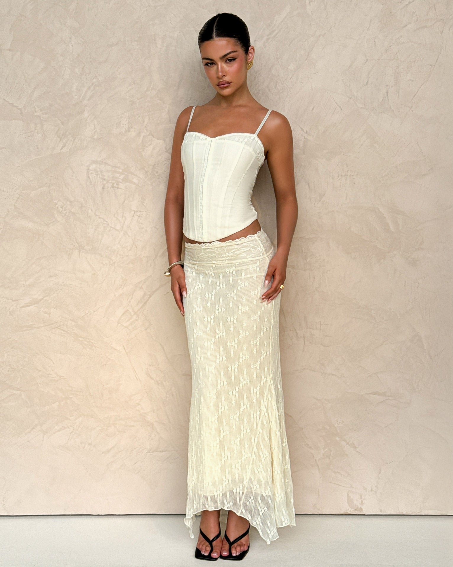A woman stands against a beige wall wearing TIGERMIST's ANTONELLA MIDI SKIRT in cream lace and white, paired with a fitted cream corset top. Her dark hair is pulled back, she wears black sandals, and maintains a neutral expression.