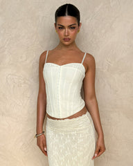 A woman in the TIGERMIST ANTONELLA TOP - WHITE : BONE : CREAM stands against a textured beige wall, wearing a fitted suiting fabric corset and a sheer lace-patterned skirt, looking directly at the camera with a neutral expression.