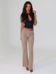 A woman with long hair stands against a neutral background, showcasing a stylish full-length silhouette. She is wearing a sleeveless black top with a keyhole cutout and the VESPER PANT in tan from TIGERMIST, featuring a low-rise waist. Black heels complete her look as one hand rests on her hair while the other hangs by her side.
