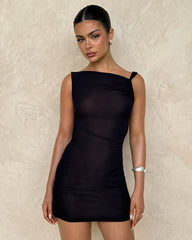 A woman with dark hair tied back wears the TIGERMIST FREYA DRESS - BLACK, a fitted mini dress with an asymmetrical neckline, paired with a silver bracelet. She stands against a textured beige wall, looking at the camera with a neutral expression.