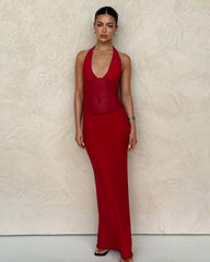 A woman with dark hair styled back wears the TIGERMIST BRESILY MAXI DRESS in red, featuring a form-fitting silhouette, halter neckline, and subtle embroidery. She stands against a light textured wall, looking at the camera neutrally.