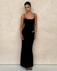 A woman stands against a textured beige wall in the TIGERMIST UMA DRESS - BLACK, a bodycon maxi with adjustable straps and side cut-out. She pairs it with black sandals and silver earrings, her hair slicked back and expression neutral.