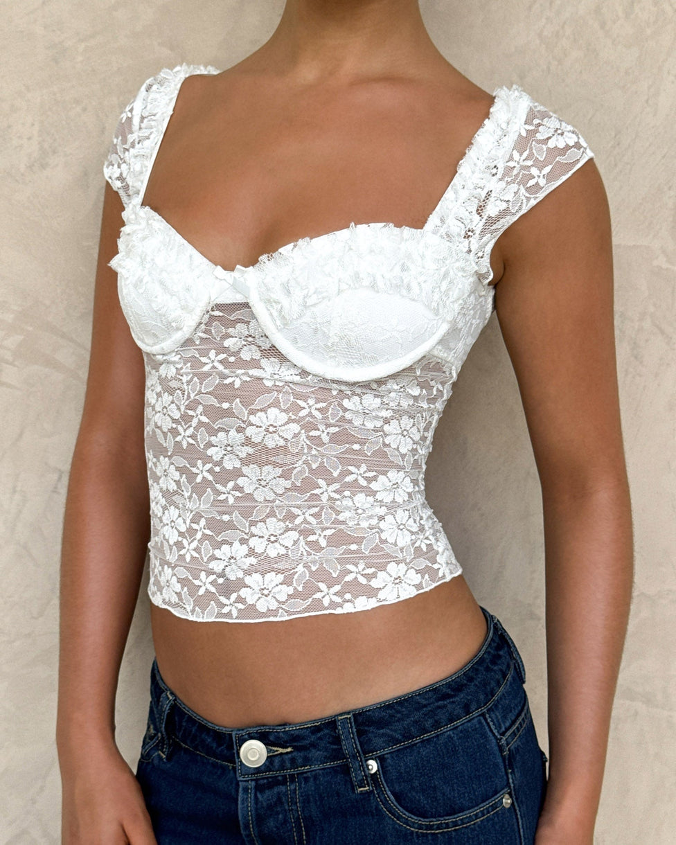 Wearing the TIGERMIST NYRA TOP - WHITE, featuring sheer shoulder straps and floral lace details, paired with blue jeans and set against a light beige textured background.