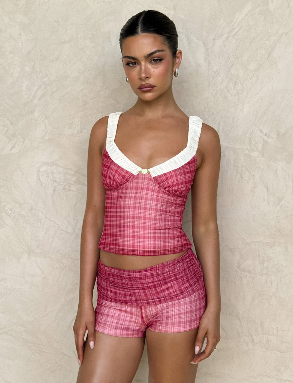 A person stands against a textured beige wall wearing a TIGERMIST AMARAN TOP - RED CHECK, featuring a white ruffled neck detail. They have slicked-back hair, hoop earrings, and a calm expression.