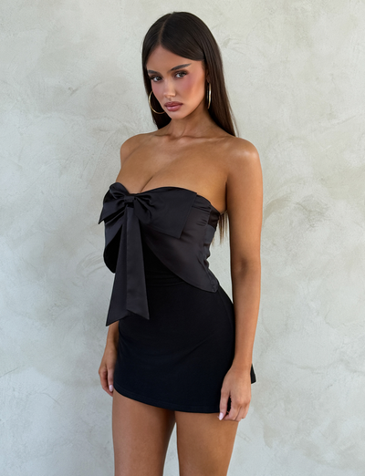 A woman with long straight hair wears the TIGERMIST GROVE MINI DRESS - BLACK, a strapless mini dress with a large satin bow on the chest. She stands before a light textured wall, looking at the camera with a neutral expression.