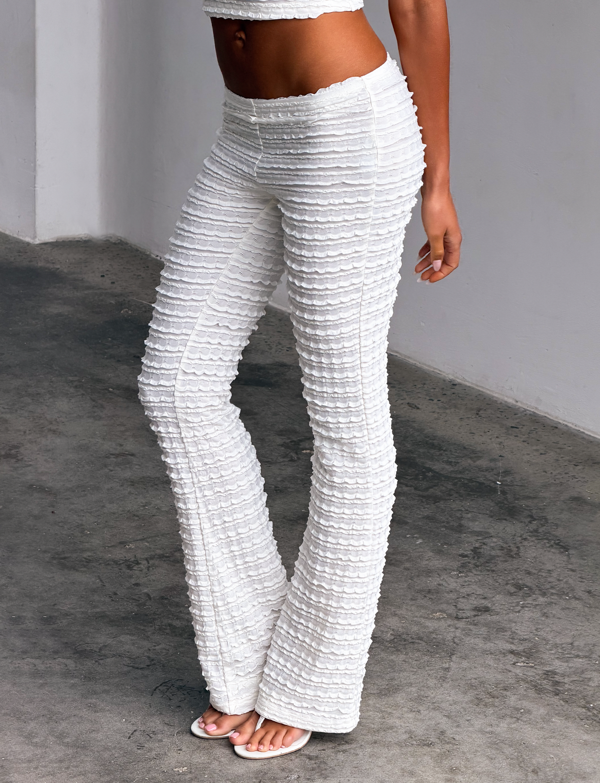 A woman stands indoors wearing the TIGERMIST ZORA PANT in cream, featuring ruffle detailing and paired with heeled sandals. Her torso is partially visible against plain white walls and a concrete floor.